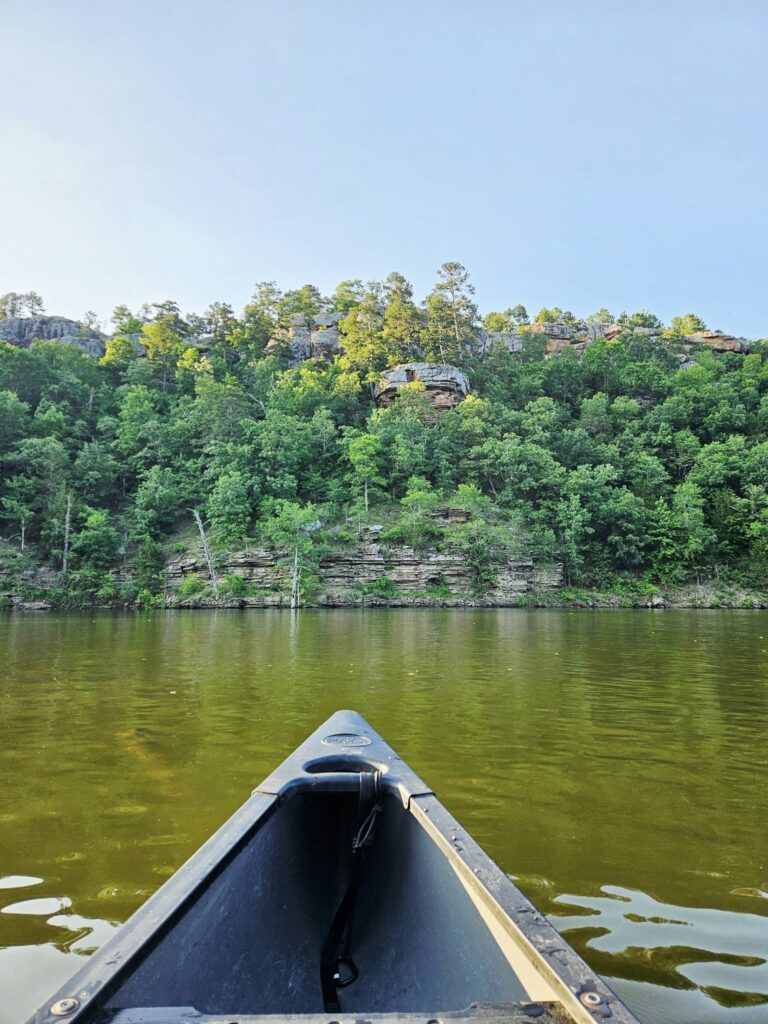 A canoe floats on a calm, greenish river facing dense, rocky forested cliffs under a clear blue sky—an inviting scene for those seeking serene kayak tours.