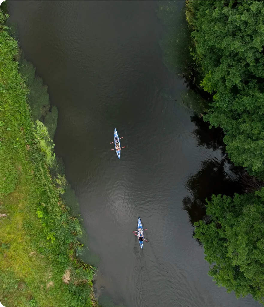 Aerial view of two people enjoying kayak tours on a narrow river, bordered by lush green vegetation on one side and dense trees on the other.
