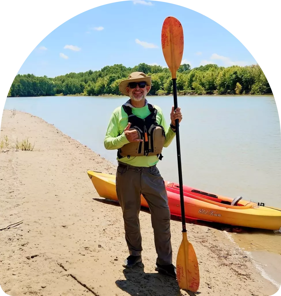 Joey, wearing outdoor clothing, stands on a sandy riverbank holding a kayak paddle, with a kayak and the river in the background.